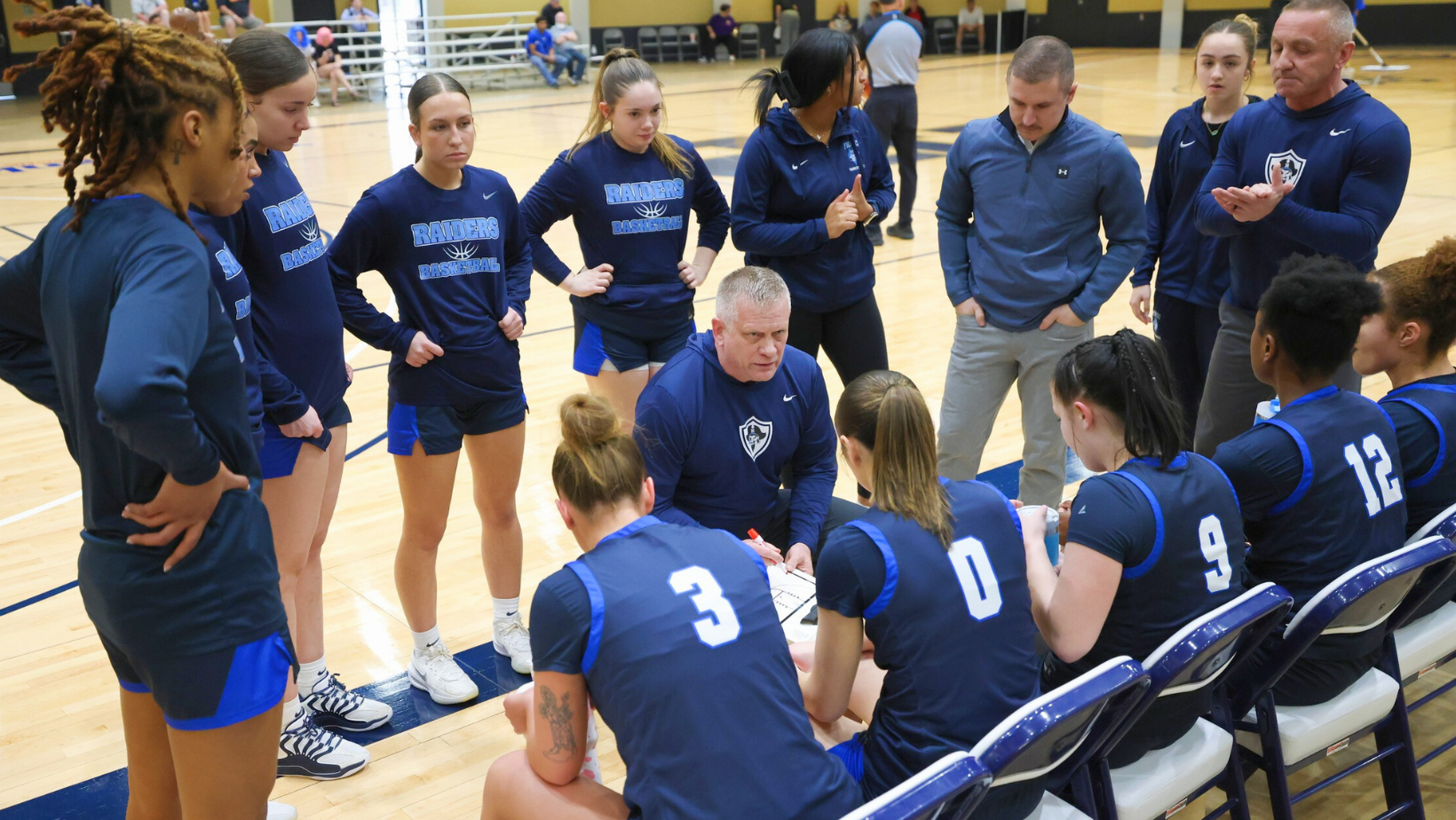 Head Coach Kevin Jones talks to his team during a timeout in the National Tournament Semi-Final Game. Girls are in navy blue uniforms sitting on bench looking at coach. Non starters are surrounding looking in as well.